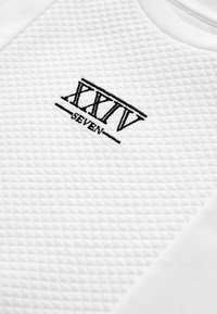 Hvidt stof med en quiltet tekstur, med et broderet logo, der læser "XXIV SEVEN" i sort, placeret centralt.