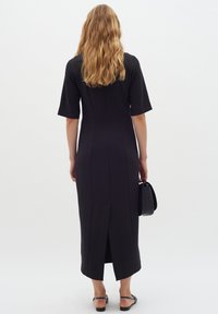 Svart knälång klänning med korta ärmar, som har en slits bak och sömlös design. Modellen håller en liten svart handväska.
