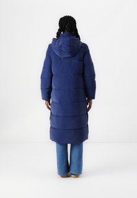 Palton lung, de culoare bleumarin, umplut cu puf, cu glugă, având secțiuni orizontale quilted și fante laterale. Purtați peste blugi albaștri și încălțăminte ușoară.