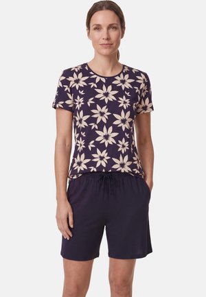 Vrouw draagt een donkerblauw T-shirt met korte mouwen met grote beige bloemenprint en effen donkerblauwe shorts met trekkoorden, staat met handen in de zakken.