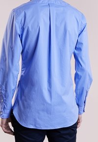 Ljusblå långärmad skjorta med en plym bak, knappade manschetter och rundad nederkant, tillverkad av ett mjukt tyg.