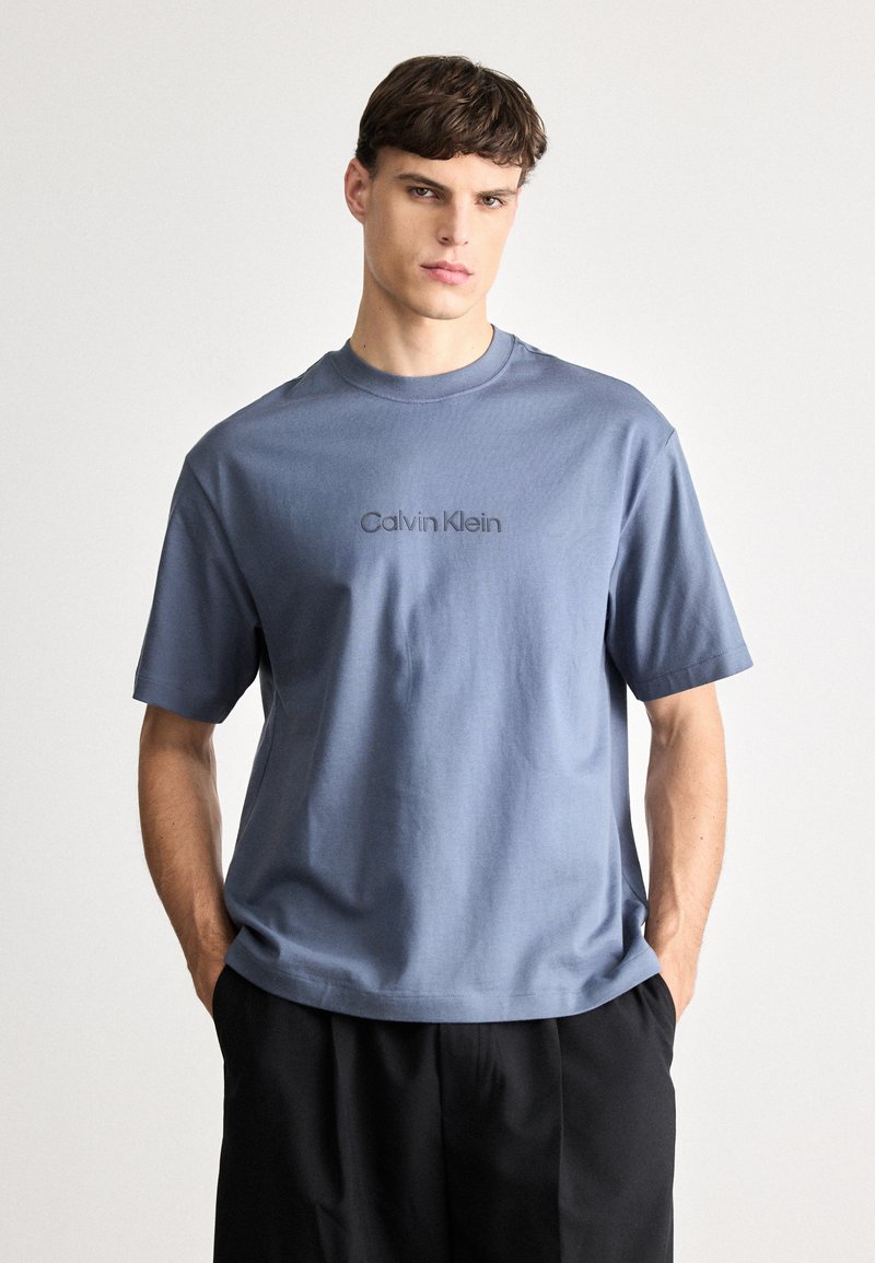 Svijetloplava pamučna majica kratkih rukava s okruglim izrezom, na prednjoj strani sivi logotip "Calvin Klein" otisnut sitotiskom.