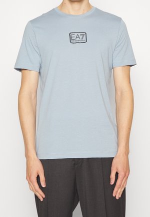 Hombre vistiendo una camiseta de manga corta azul claro con un pequeño parche negro con el logo que dice "EA7 EMPORIO ARMANI" en el pecho.