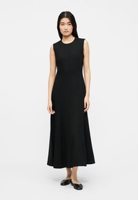 Naine seisab, kandes varrukateta musta pahkluu pikkust kleiti ja musti madalaid kingi hallil ühtlasel taustal.