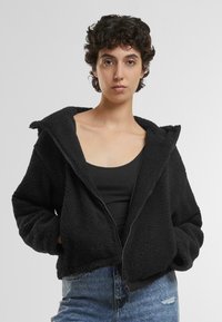 Svart fleece zip-up-jacka med bred huva, kort design och texturerad yta. I kombination med en svart linne och blå jeans.