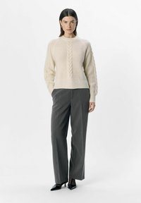 Cremefarvet sweater med et tekstureret kabeldesign, parret med grå brede bukser og sorte spidse hæle. Glat strik, afslappet pasform.