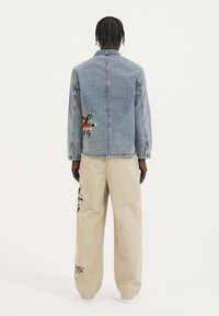 Denimtakki, jossa on brodeerattu graafinen kuva selässä, yhdistettynä beigekatsoihin, joissa on samanlainen brodeeraus. Molemmat ovat rentoja ja oversized-mallisia.