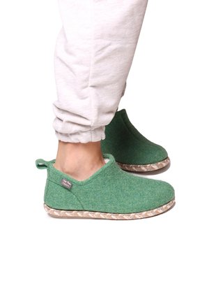 Feet wearing green wool slippers with braided soles and beige jogger pants on a white background.