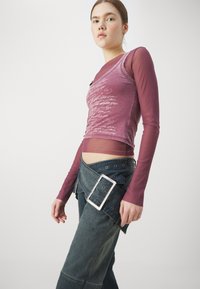 Langermet bordeaux topp lagd over en tettsittende rosa tank, parret med blå denimbukser med hull detalj og en stor sølvspenne.