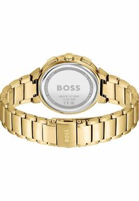 Orologio in acciaio inossidabile dorato con cassa tondeggiante, bracciale in metallo e finitura lucida. Il retro mostra il logo del marchio e i dettagli sulla resistenza all'acqua.