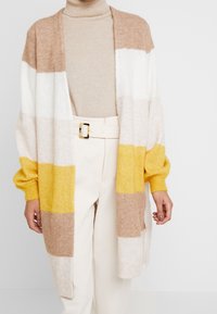 Kardigan sięgający kolan w pastelowe paski w kolorze żółtym, białym i beżowym. Miękka, dziergana faktura z szerokimi rękawami i otwartym przodem.