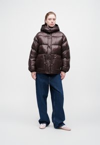 Chaqueta acolchada marrón con capucha, que presenta bafles sobredimensionados y dos bolsillos frontales, combinada con vaqueros azules holgados y zapatos ligeros.