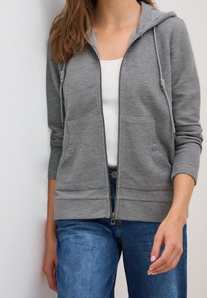 Femme portant un sweat à capuche gris zippé avec un t-shirt blanc en dessous et un jean bleu, debout devant un fond blanc.