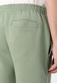 Jogging en coton vert avec une taille élastique, présentant une petite poche arrière et une finition lisse et texturée. Poche latérale visible.