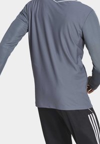 Personne portant une chemise en tissu texturé à manches longues grise et un pantalon noir avec des rayures blanches sur les côtés, de dos avec un bras tendu.