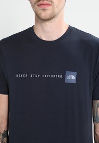 T-paita tummansinisestä puuvillasta, jossa on valkoisella teksti "ÄLÄ KOSKAAN PYSÄHDY TIETÄMÄÄN" ja pieni logopatch rintamuksessa. Lyhyet hihat.