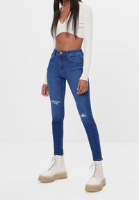Jeans ajustados de mezclilla azul de cintura alta con detalles desgastados, combinados con un top blanco de manga larga cropped y botas de plataforma color crema.