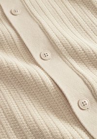 Beige cardigan med texturerad ribbning, som har tre runda knappar. Tyget är mjukt och vävt med ett subtilt mönster.