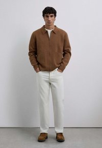 Mand står med hænderne i lommerne iført en brun zip-up trøje, hvide bukser og brune ruskindsko foran en ensfarvet grå baggrund.