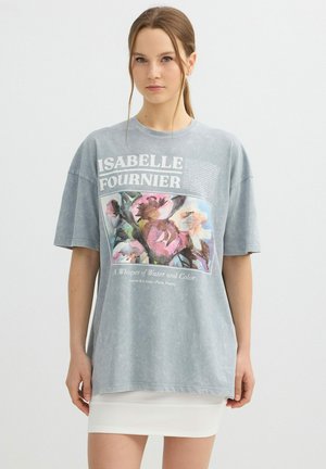 Joven mujer con camiseta de gran tamaño desgastada azul grisácea con estampado floral y texto "Isabelle Fournier" combinada con falda blanca.