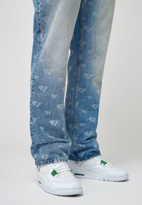 Ljust blå denimjeans med ett upprepat logomönster, tillsammans med vita sneakers med gröna detaljer och meshnätpaneler.