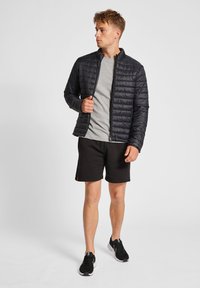 Svart vadderad jacka med hög krage och dragkedja, bärs över en ljusgrå t-shirt, ihop med svarta shorts och träningsskor.