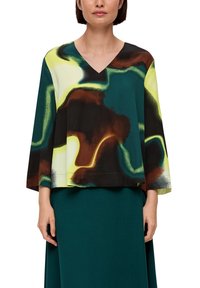 Vrouw in een meerkleurige blouse met abstracte print en V-hals met brede mouwen, gecombineerd met een donkergroene rok, staand tegen een witte achtergrond.
