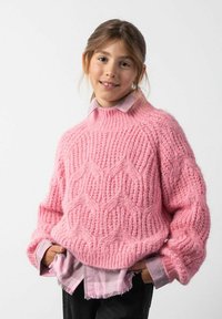 Różowy sweter dziany z teksturowanym wzorem w zygzak, wysoka szyja, luźny krój. Nosi bardzo jasną różową koszulę z kołnierzykiem. Czarnie spodnie.