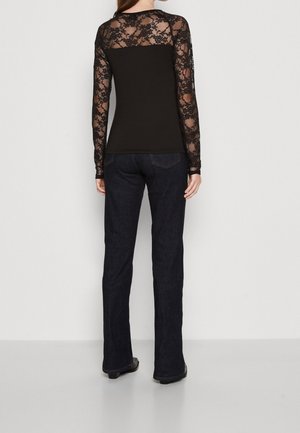 Femme portant un haut en dentelle noire à manches longues et un jean foncé, debout face à un mur blanc uni, mains détendues le long du corps.