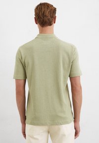 Chemise polo verte claire à manches courtes en tissu doux, avec un col classique et une texture subtile. L'ourlet latéral présente une petite étiquette.