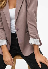 Upravený blazer z měkkého fialového materiálu, s dvojicí fialových knoflíků, kapsou a vyhrnutými pruhovanými manžetami. Součástí jsou černé kalhoty a dřevěná stolička.