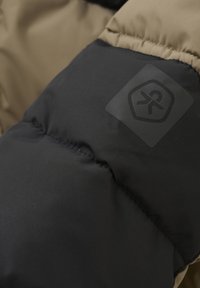 Juoda ir šviesiai ruda pūkinės striukės rankovė, pasižyminti lygiu paviršiumi ir subtiliu logotipo užtrauktuku. Drabužis yra izoliuotas šilumai.