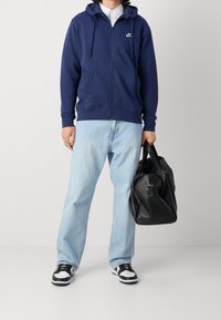 Person bär en marinblå Nike-hoodie med dragkedja, ljusblå jeans, svartvita sneakers och håller en svart läderduffelväska mot en enfärgad bakgrund.