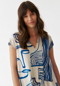 V-hals jurk met cap sleeves in lichte stof, met blauwe abstracte patronen op een beige achtergrond. Opvallende lijnen en vormen accentueren het ontwerp.