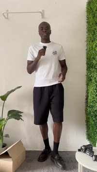 Vit t-shirt med logotyp, svarta shorts, svarta skor. Rummet har en grön växt och en vit vägg med en strukturerad yta och ett litet bord.