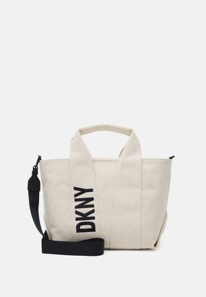 Sac cabas en toile beige avec deux poignées, une bandoulière noire amovible et un texte noir audacieux "DKNY" sur la poignée avant.