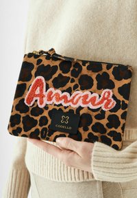 Hand die een clutch met luipaardprint vasthoudt, met "Amour" geborduurd in rood en roze, en een zwart Codello-label op een beige gebreid kledingstuk.
