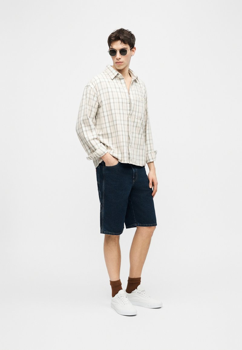 Giovane uomo con occhiali da sole rotondi, camicia beige a quadri larga, shorts in denim scuro, calzini marroni e sneakers bianche in piedi davanti a uno sfondo semplice.