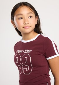 Ragazzina con lunghi capelli scuri, che indossa una maglietta marsala con la scritta "New York 89" e rifiniture bianche, che guarda leggermente verso sinistra su uno sfondo neutro.