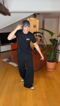 A person wears a black shirt and wide black pants, with a patterned black beanie and silver shoes, posing in a living room with plants.