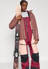 Lyžiarska výbava s hnedou bundou, ružovými a burgundskými lyžiarskymi nohavicami s čiernymi akcentmi, drží lyže s čiernymi viazaniami a vzorovaným sklzom.