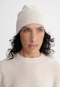 Beige gebreide beanie met een geribbelde textuur. Slouchy ontwerp, zacht materiaal en een taps toelopende bovenkant. Gedragen met een bijpassende gebreide trui.