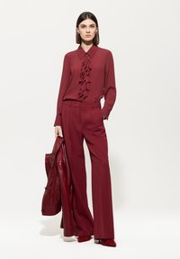 Bordeauxfarbene Bluse mit gerafftem Vorderteil, kombiniert mit weiten Hosen. Hält eine passende Lederjacke; akzentuiert durch haarige, rote Absätze.