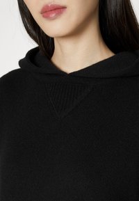 Nærbillede af en person iført en sort strikket hættetrøje, der viser den nedre del af ansigtet, nakken og de teksturerede stofdetaljer.