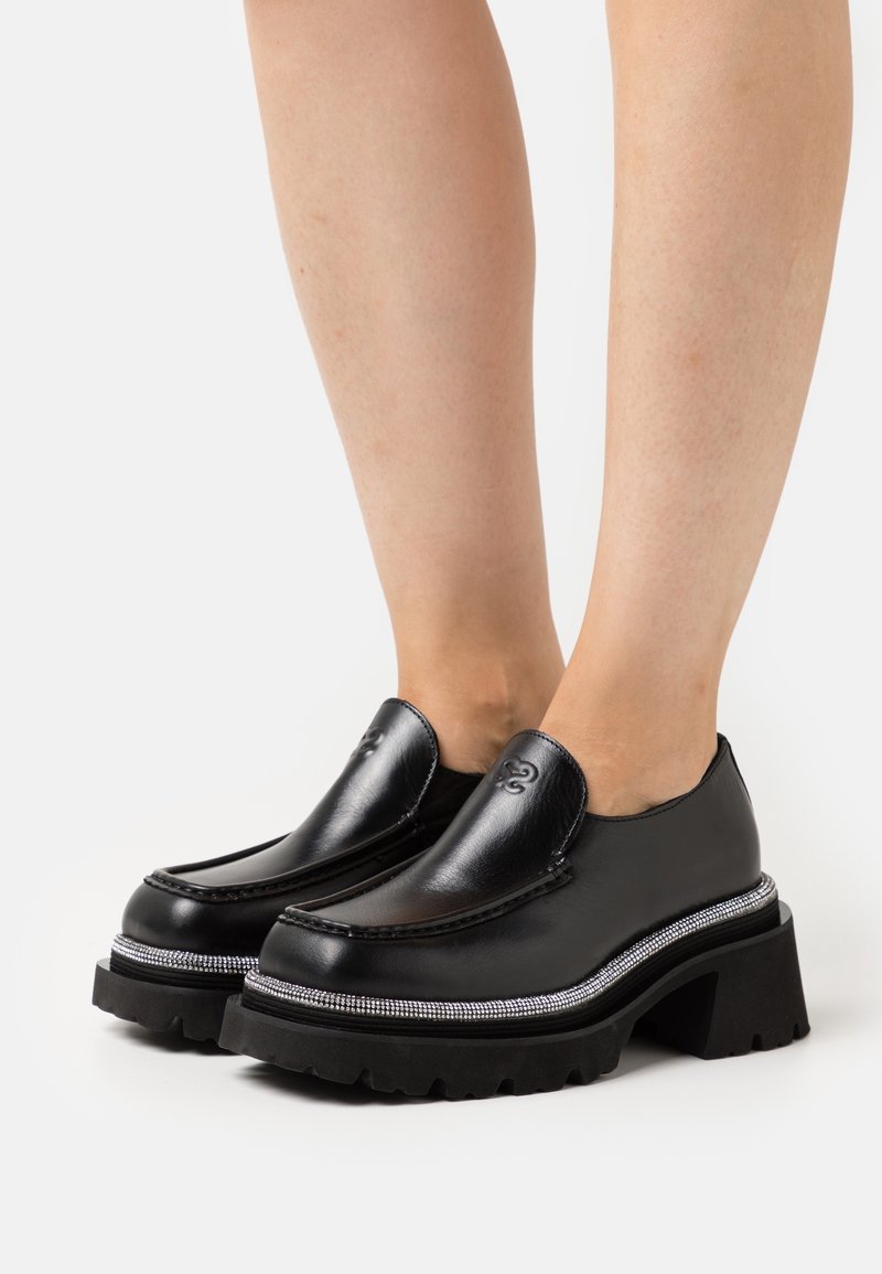 Mocassins à plateforme en cuir noir avec semelles épaisses et garniture argentée portés pieds nus sur fond blanc.