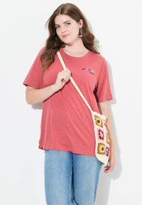 Camiseta corta de manga roja con el texto "OPTIMISTA", combinada con jeans de color azul claro y un bolso crema con patrones de ganchillo coloridos.