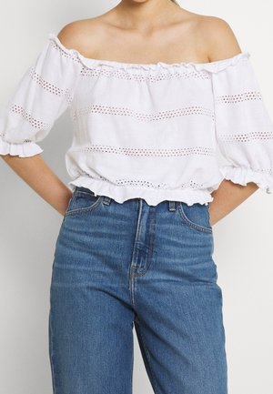 Femme portant un chemisier blanc à épaules dénudées avec des détails en œillets et un jean bleu taille haute, les mains posées sur les hanches.