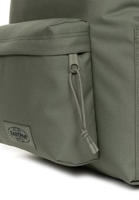 Mochila de tela verde con un gran bolsillo frontal, cordón de cierre y un parche prominente con el logo de la marca. Detalle de superficie texturizada.