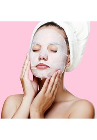 Gesichtsmaske, die auf dem Gesicht einer Frau aufgetragen ist, mit einer glatten, weißen Textur und Aussparungen für Augen, Nase und Mund, vor einem pinken Hintergrund.