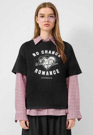 Jonge vrouw met lang haar draagt een grote bril met heldere glazen, een gelaagde roze geruite blouse en een zwart T-shirt met de tekst "No Chance Romance" en een hartontwerp.
